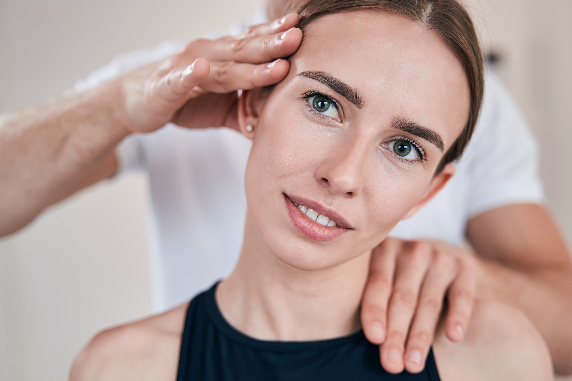 Beautiful female posing at the photo camera while unrecognized massage therapist examining her neck